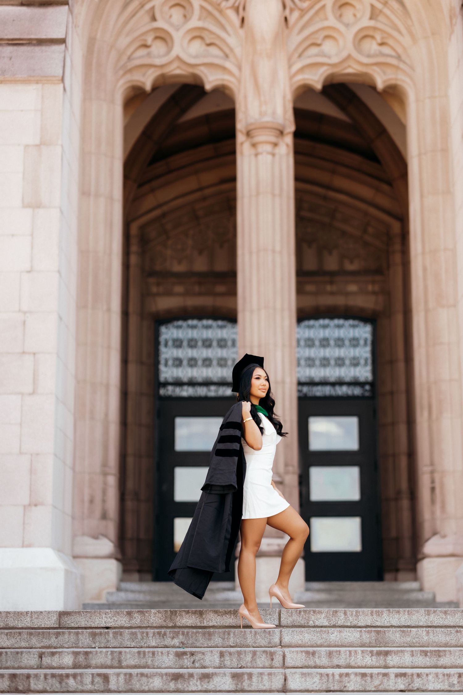 Graduate in white dress and black robe poses in Gothic archway.