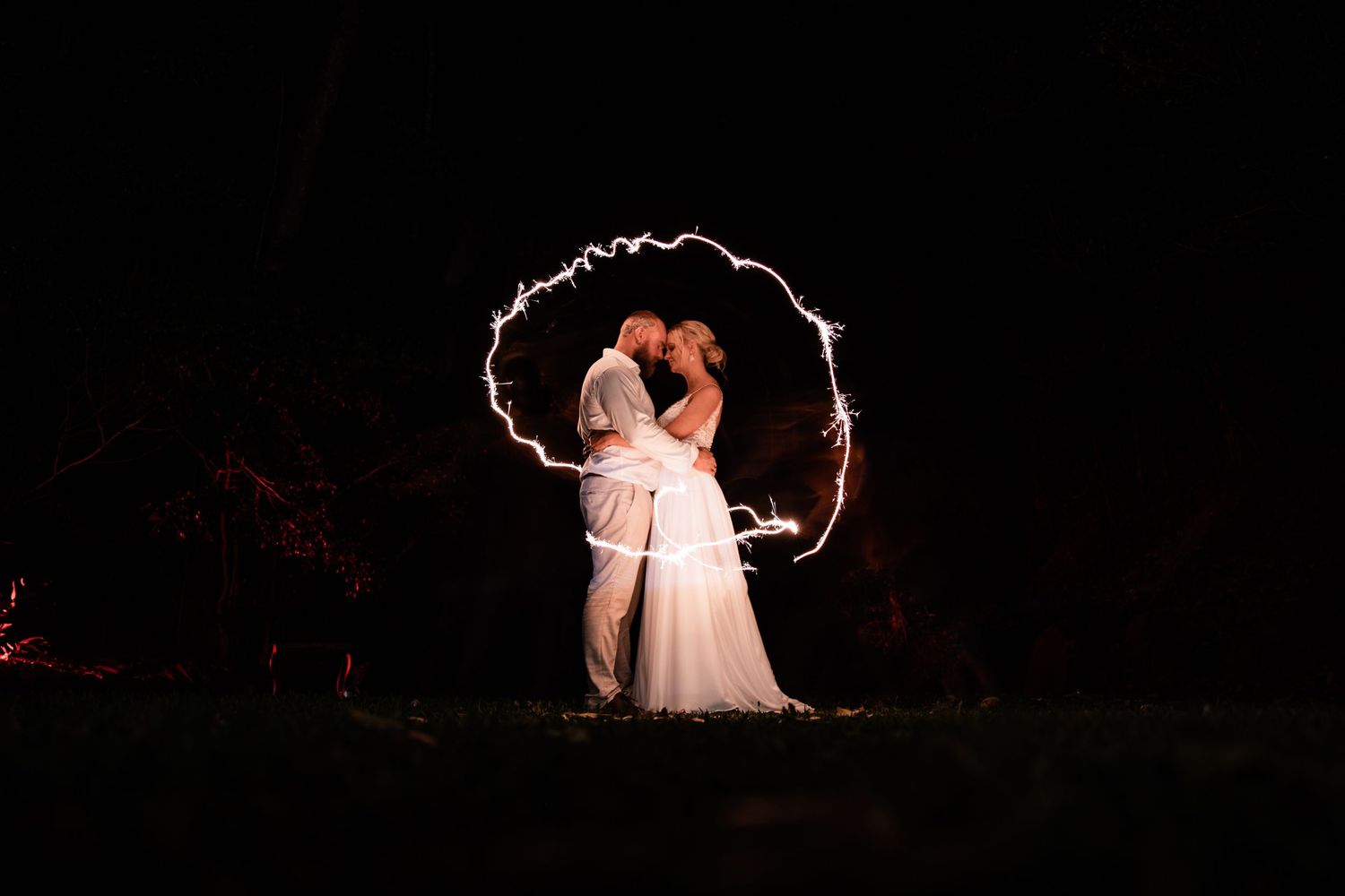 Light painting creates glowing heart shape around couple in romantic nighttime wedding photo.