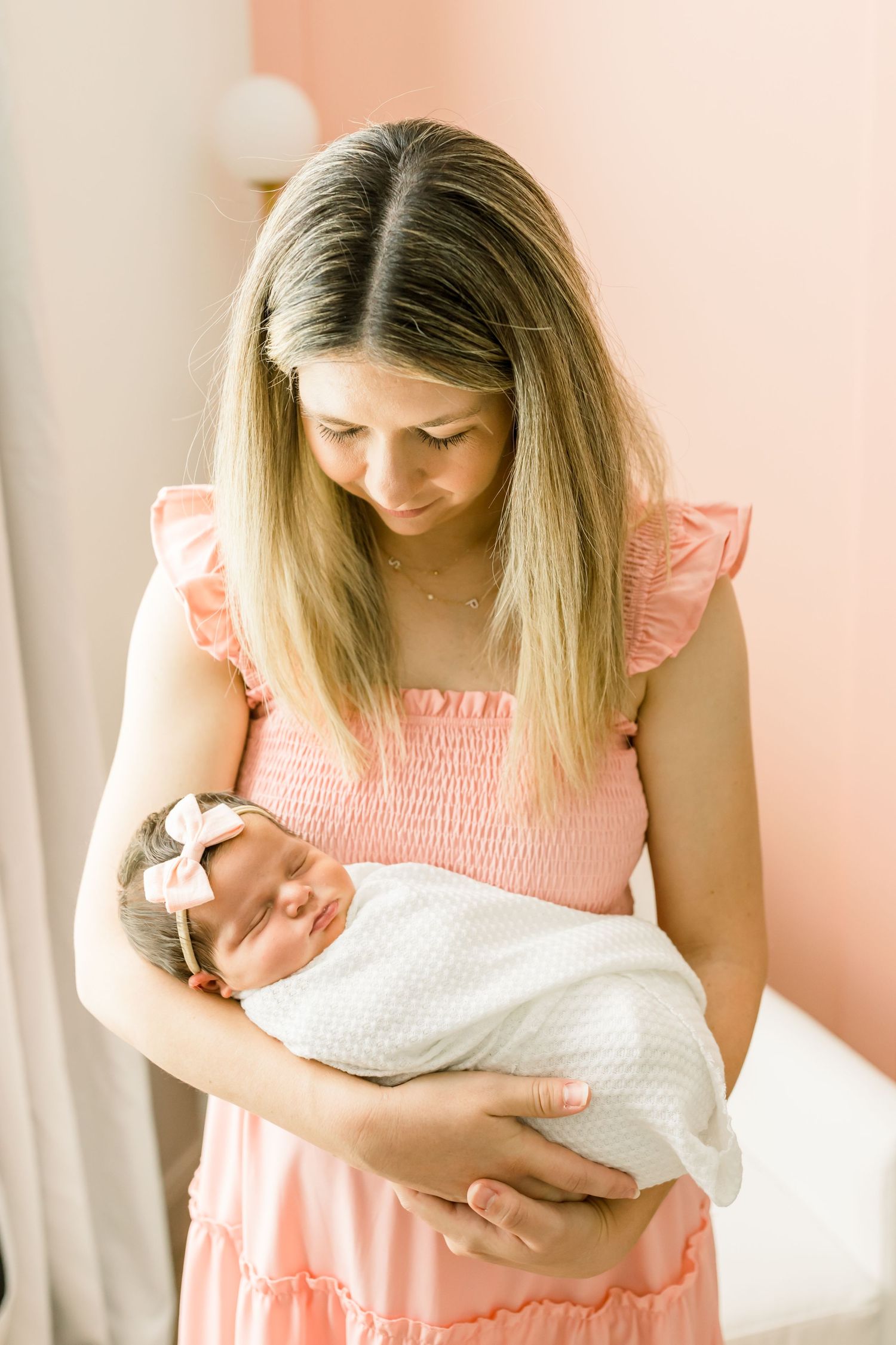 Mother in pink dress tenderly gazes at sleeping newborn.