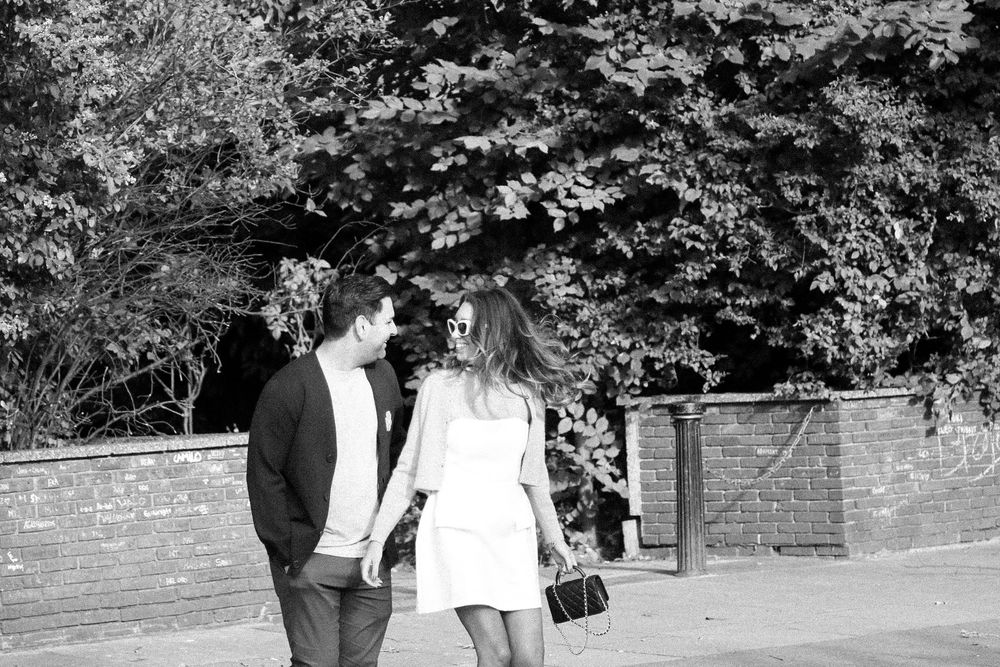 Black and white photo of couple walking across street crossing under tree canopy.