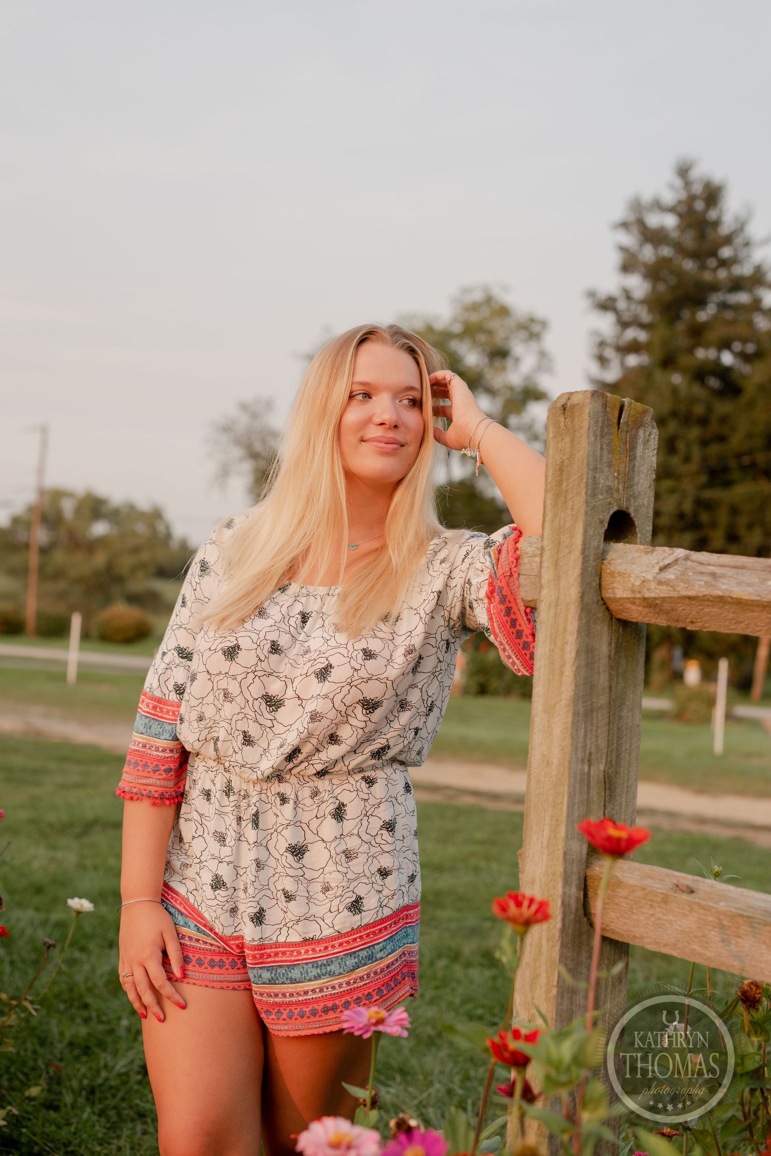 A woman poses by a rustic fence surrounded by red flowers at dusk.