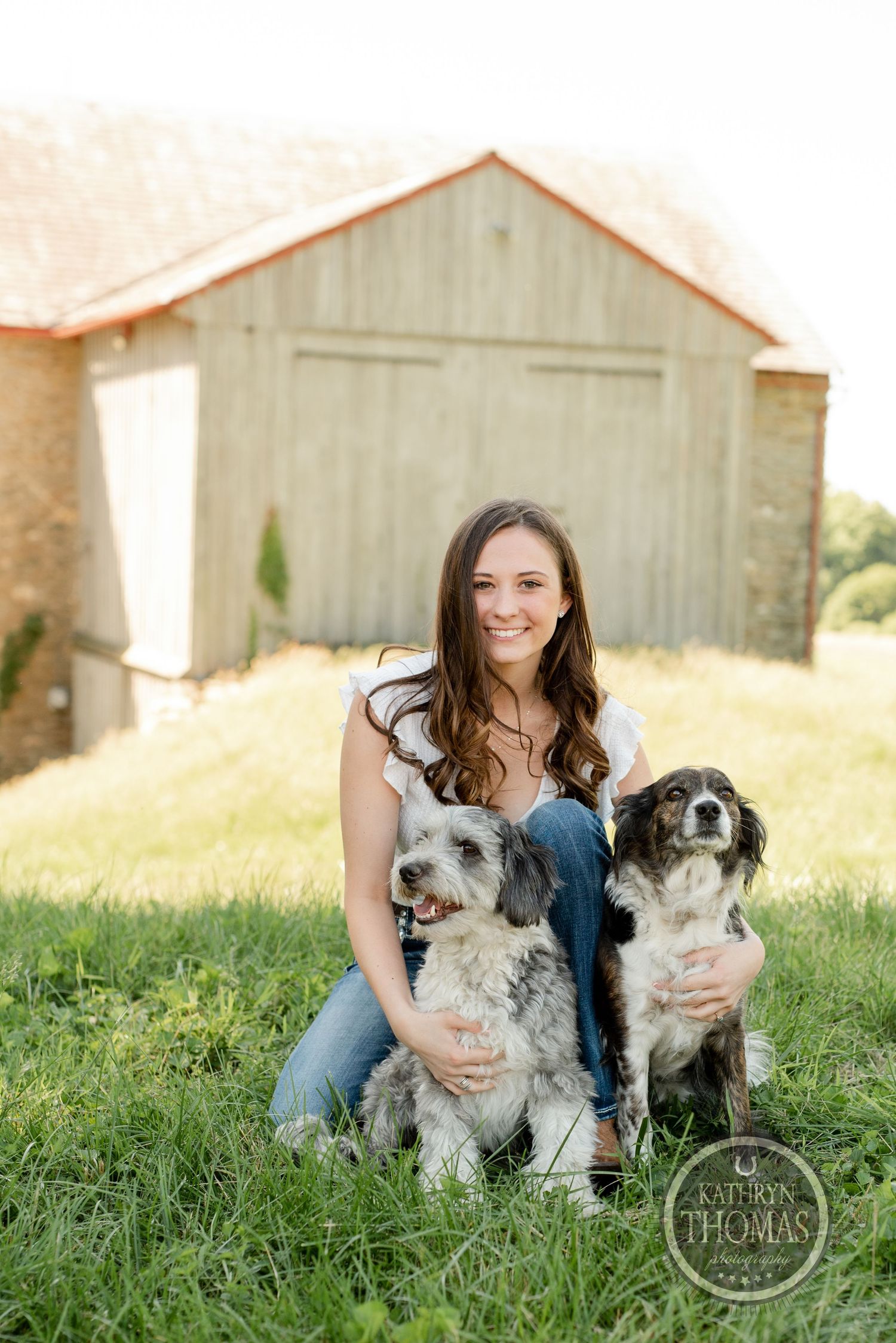 Woman poses with two friendly dogs in front of rustic barn setting.