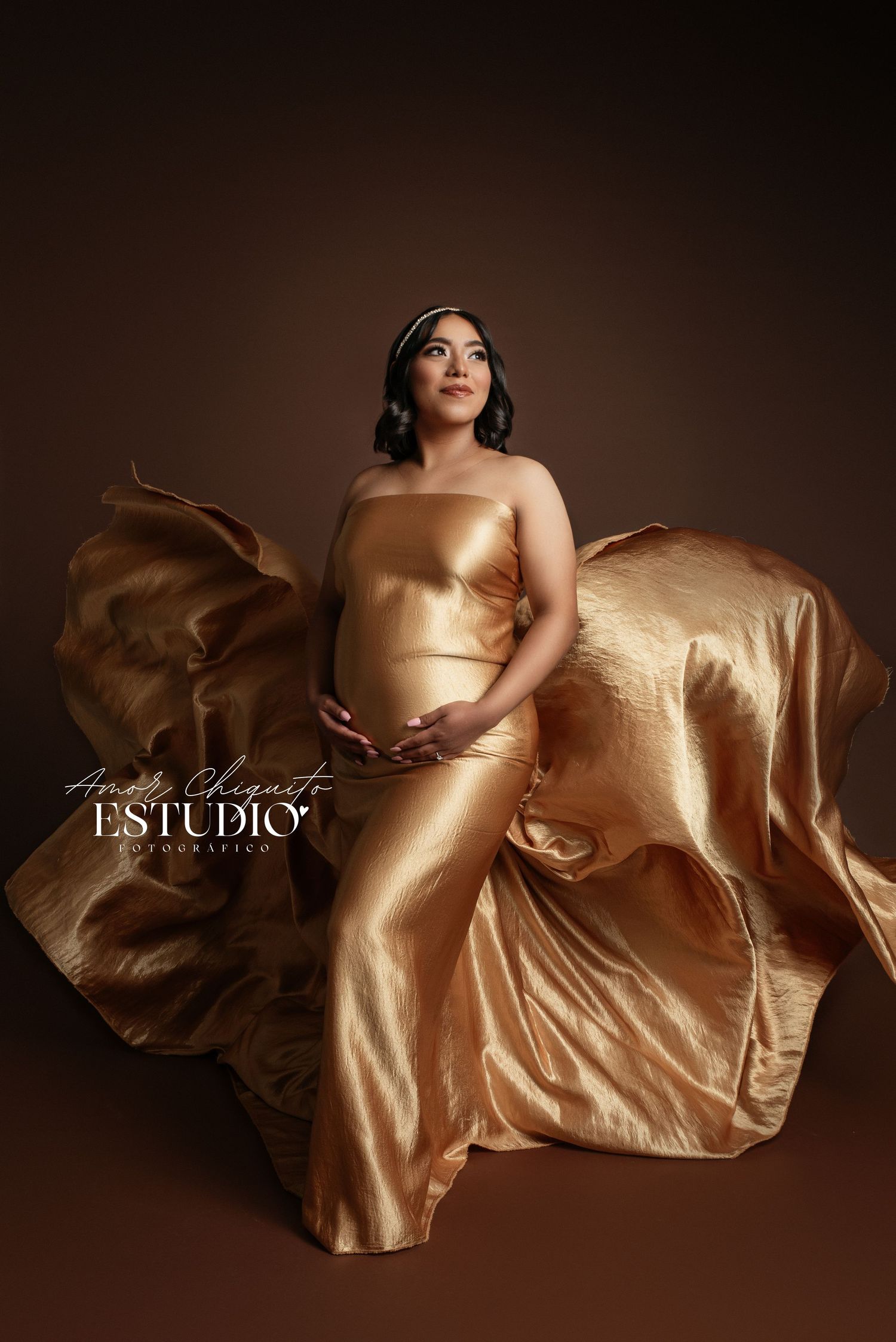 Artistic maternity photo featuring flowing gold fabric against a dark brown background.