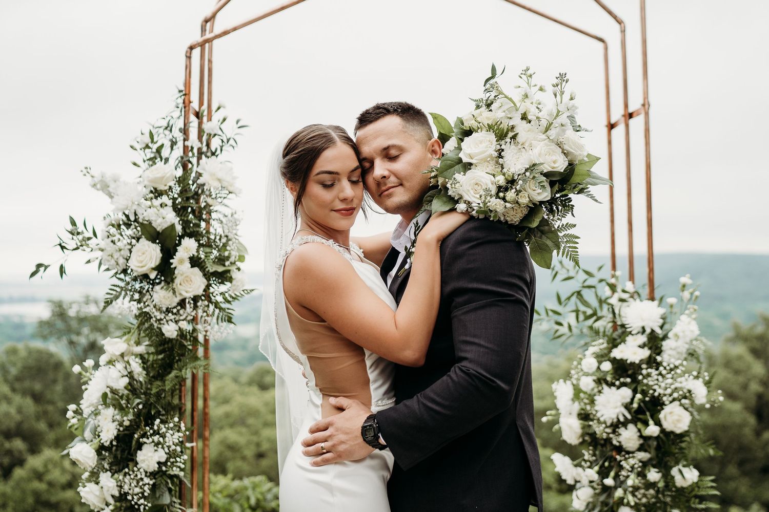 A couple embracing under a floral arch decorated with white blooms and greenery.