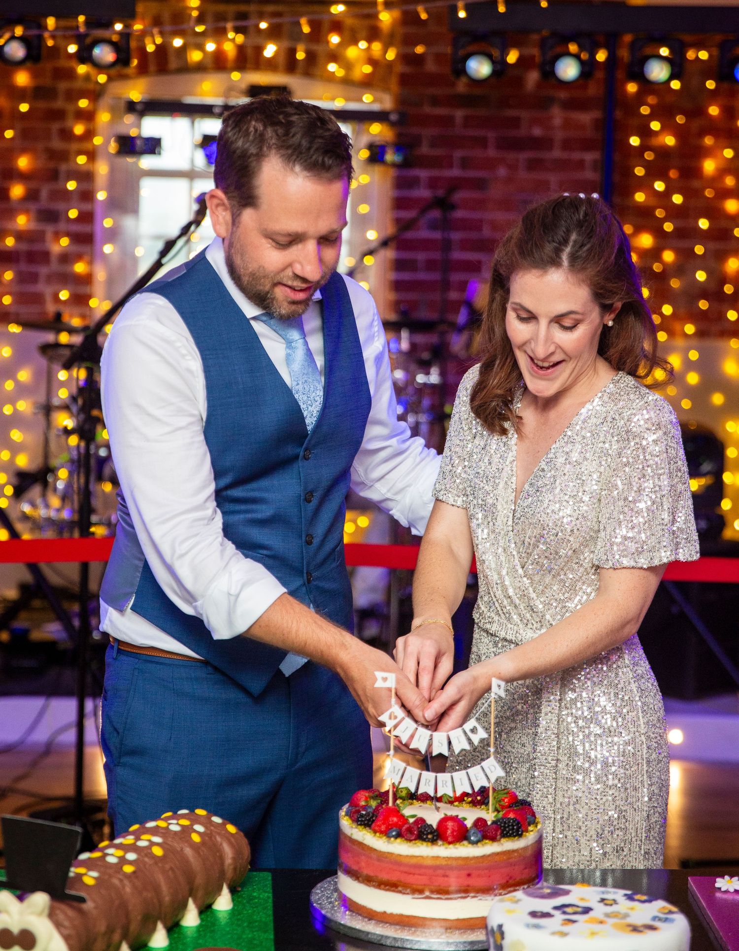 A couple in formal attire cuts their wedding cake together on a stage decorated with warm string lights.