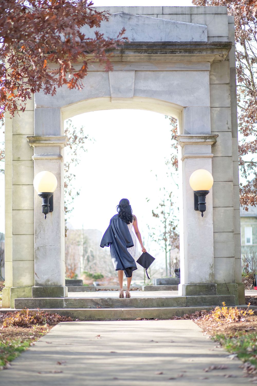 Graduate in cap and gown walks through a stone archway on a college campus in autumn.