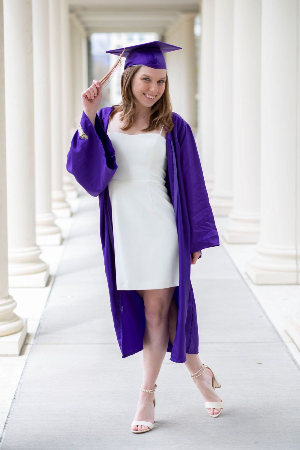 Smiling graduate in purple cap and gown poses between white columns in a white dress.