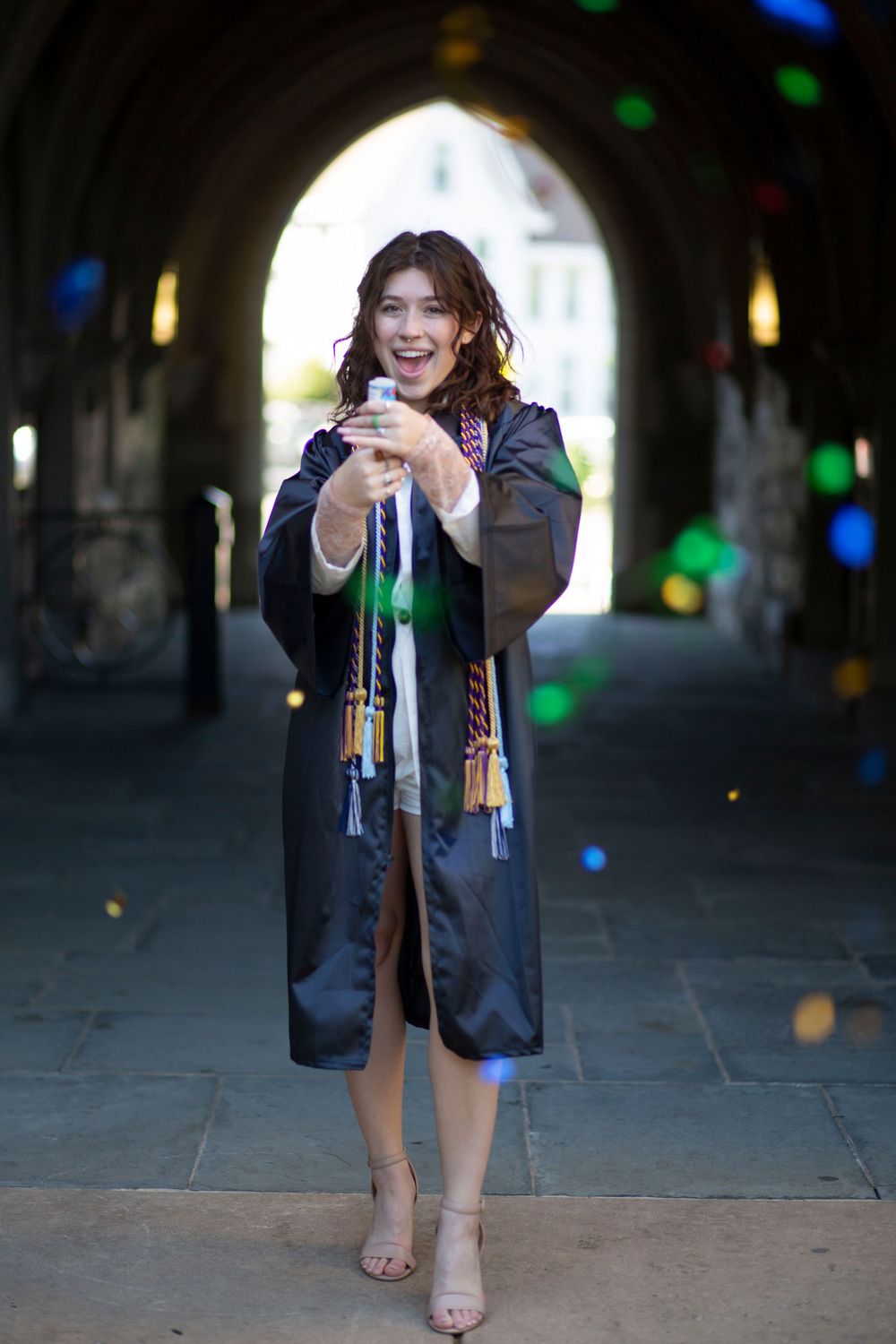 Joyful graduate in black robe holds diploma under colorful bokeh lights in stone archway.
