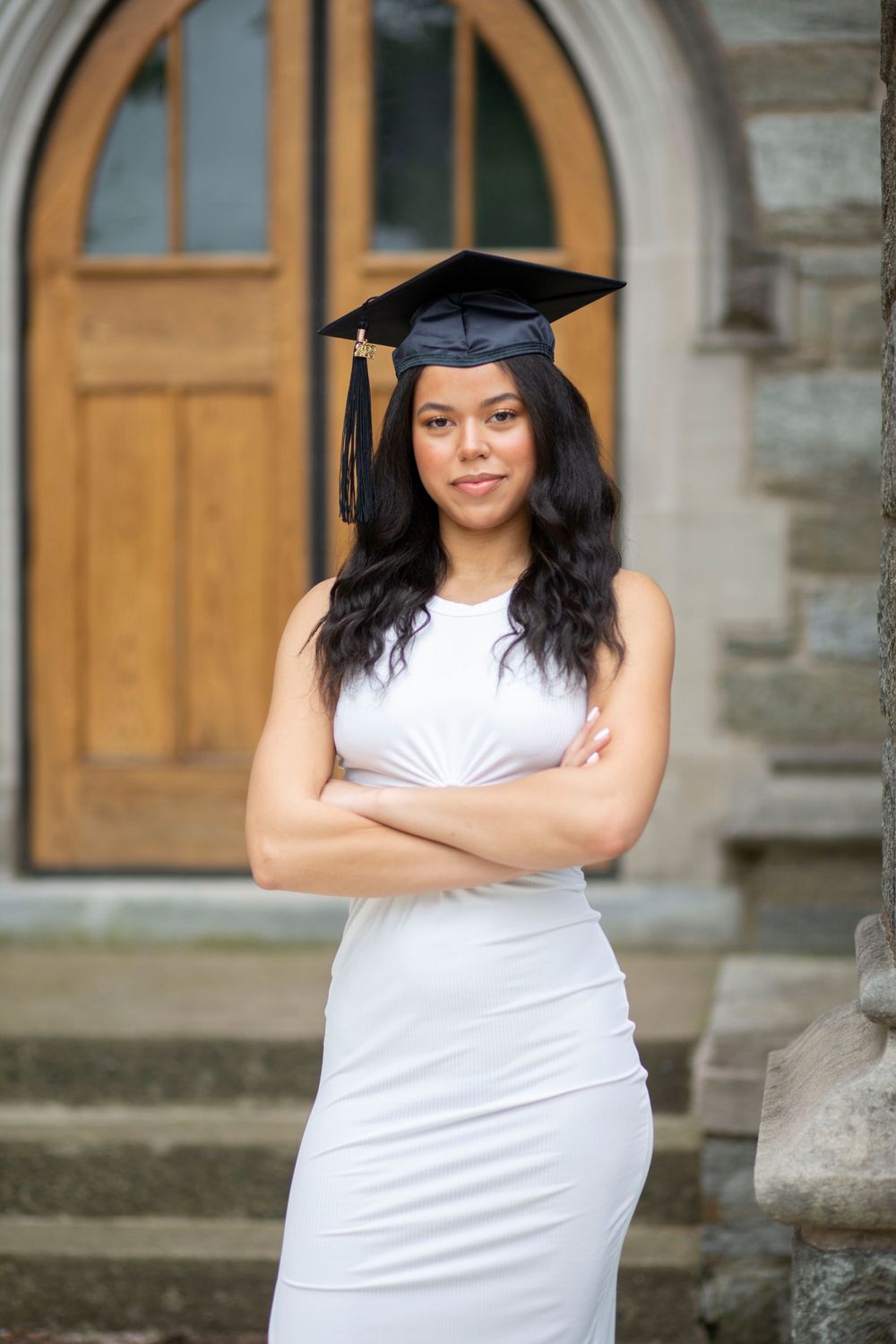 Graduate in white dress and cap poses confidently with arms crossed before arched wooden doors.