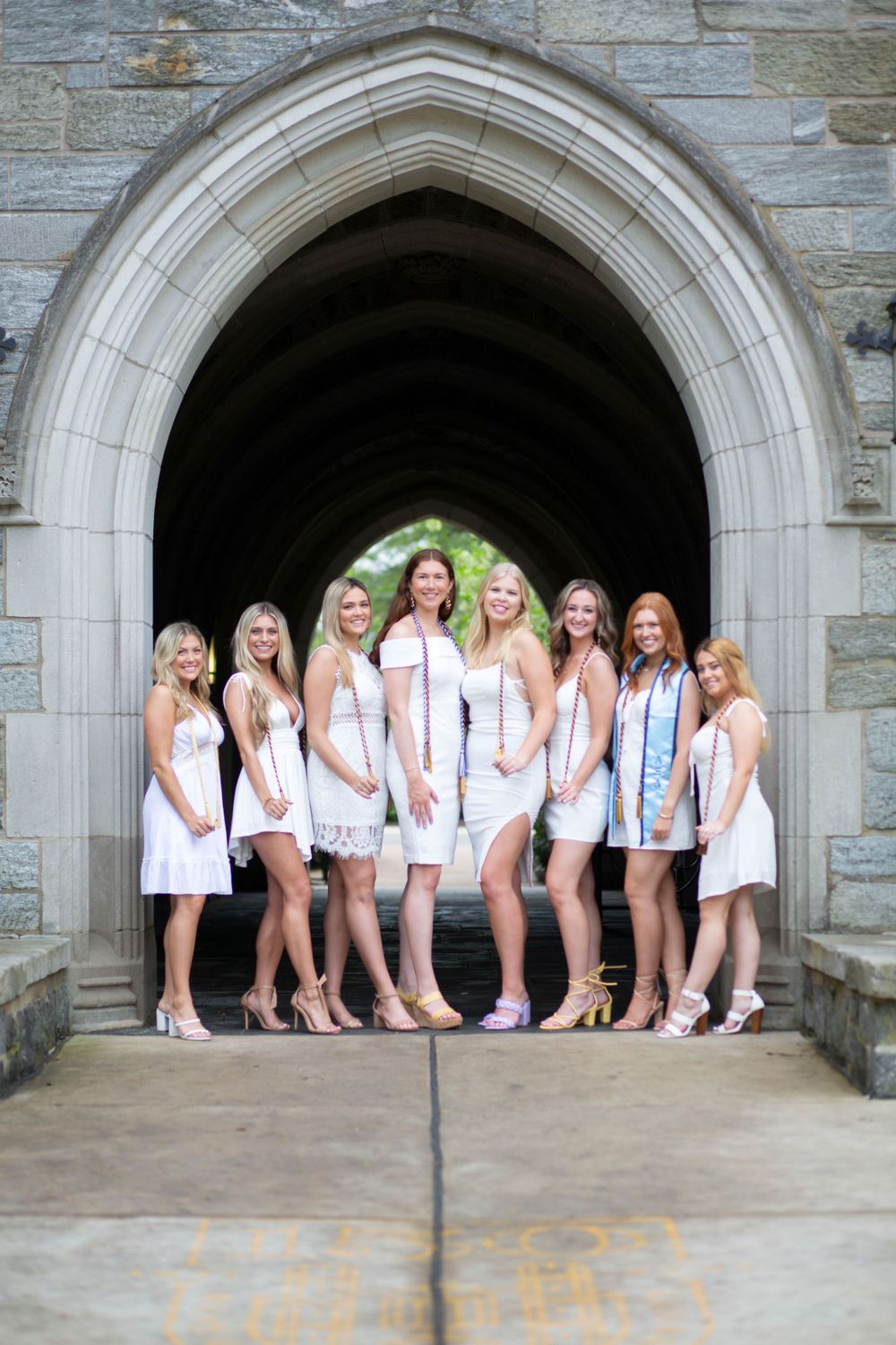 Group of eight women in white dresses pose together under Gothic stone archway at graduation.
