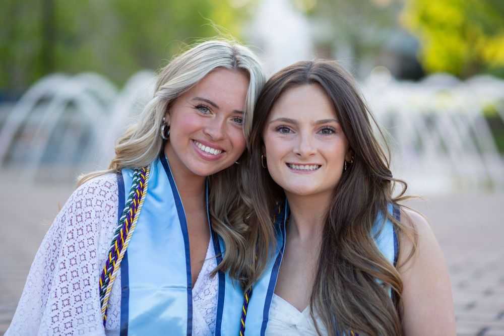 Two smiling young women in graduation stoles posing together outdoors near a fountain.