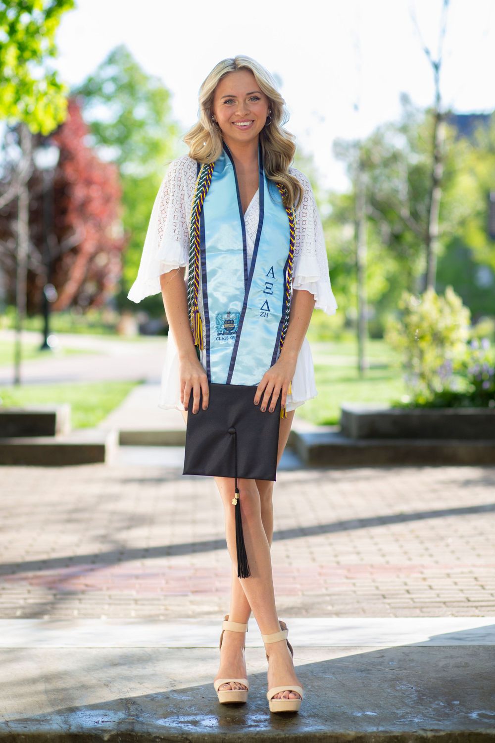 Blonde woman in white lace dress and graduation stole posing outdoors on a campus walkway.