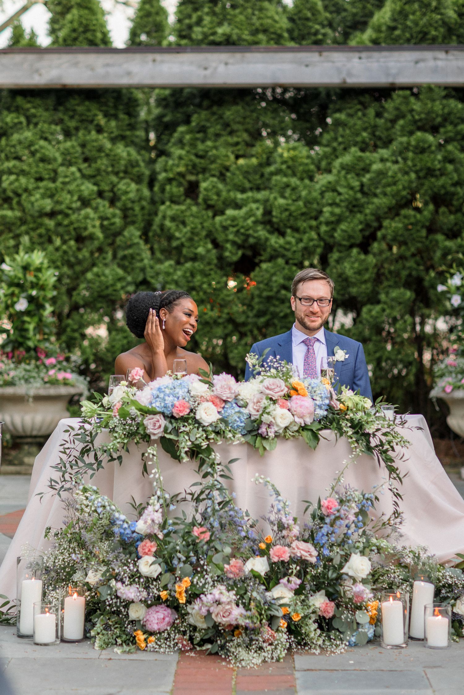 Ornate sweetheart table decorated with lush floral arrangements and candles under an outdoor pergola.