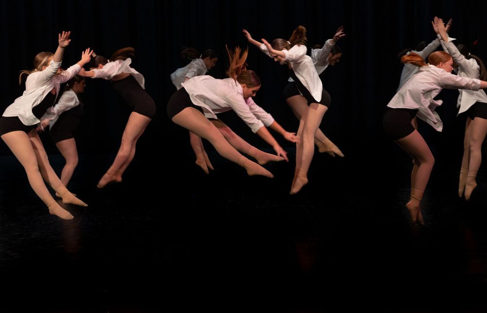 Contemporary dancers in white dresses perform leaping movements against a dark background.