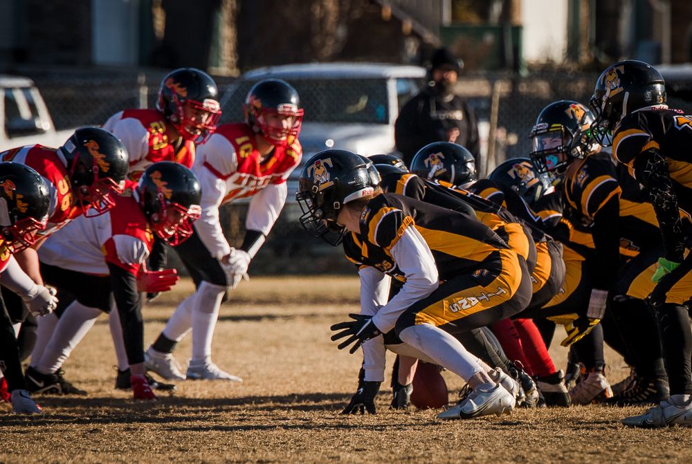 Football teams in red and black-yellow uniforms clash at line of scrimmage during sunset game.