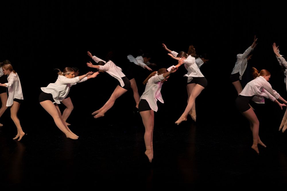 Multiple dancers in light dresses perform synchronized leaping movements across a dark stage.