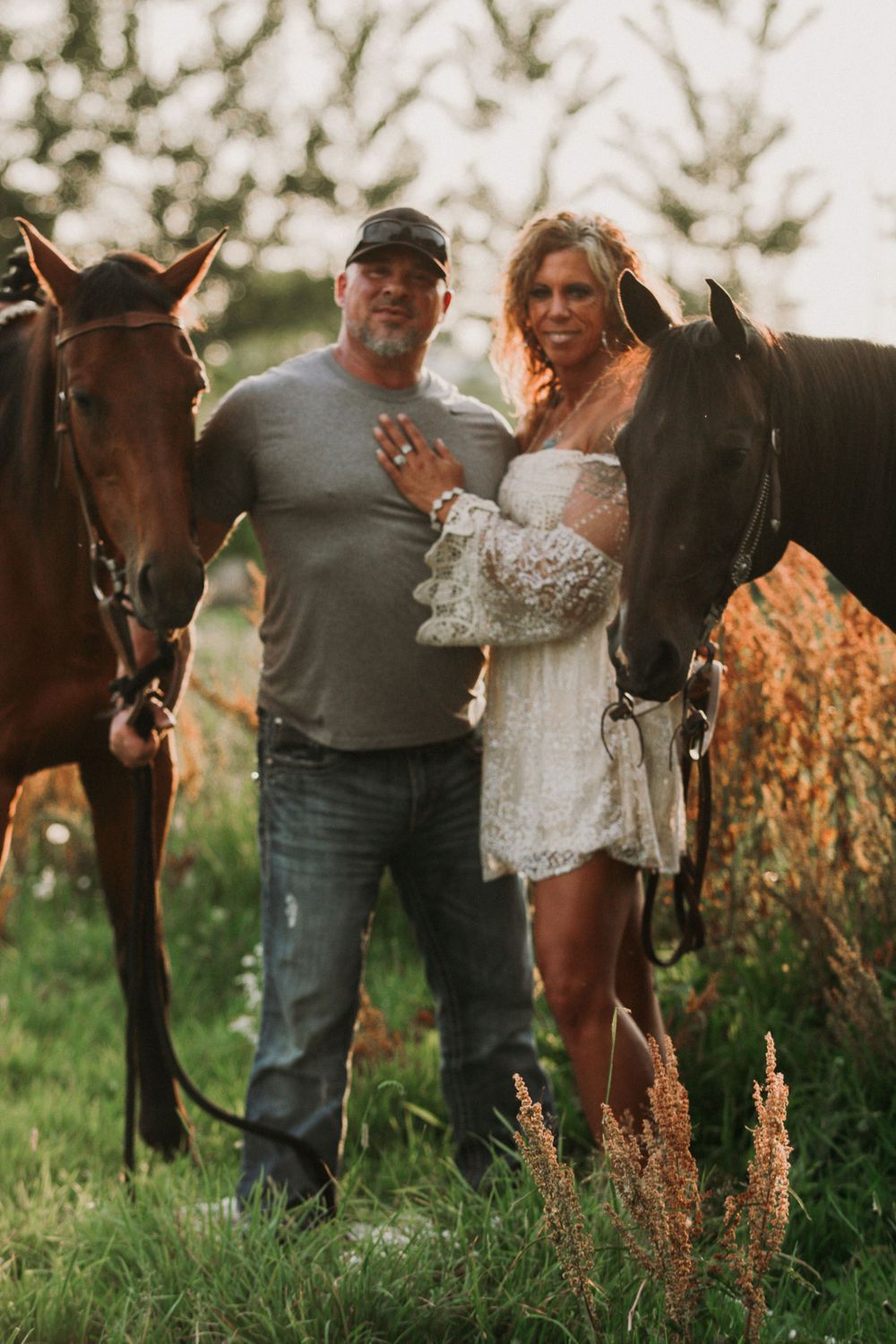 Outdoor engagement photoshoot featuring a couple posing with horses in a rustic field setting.