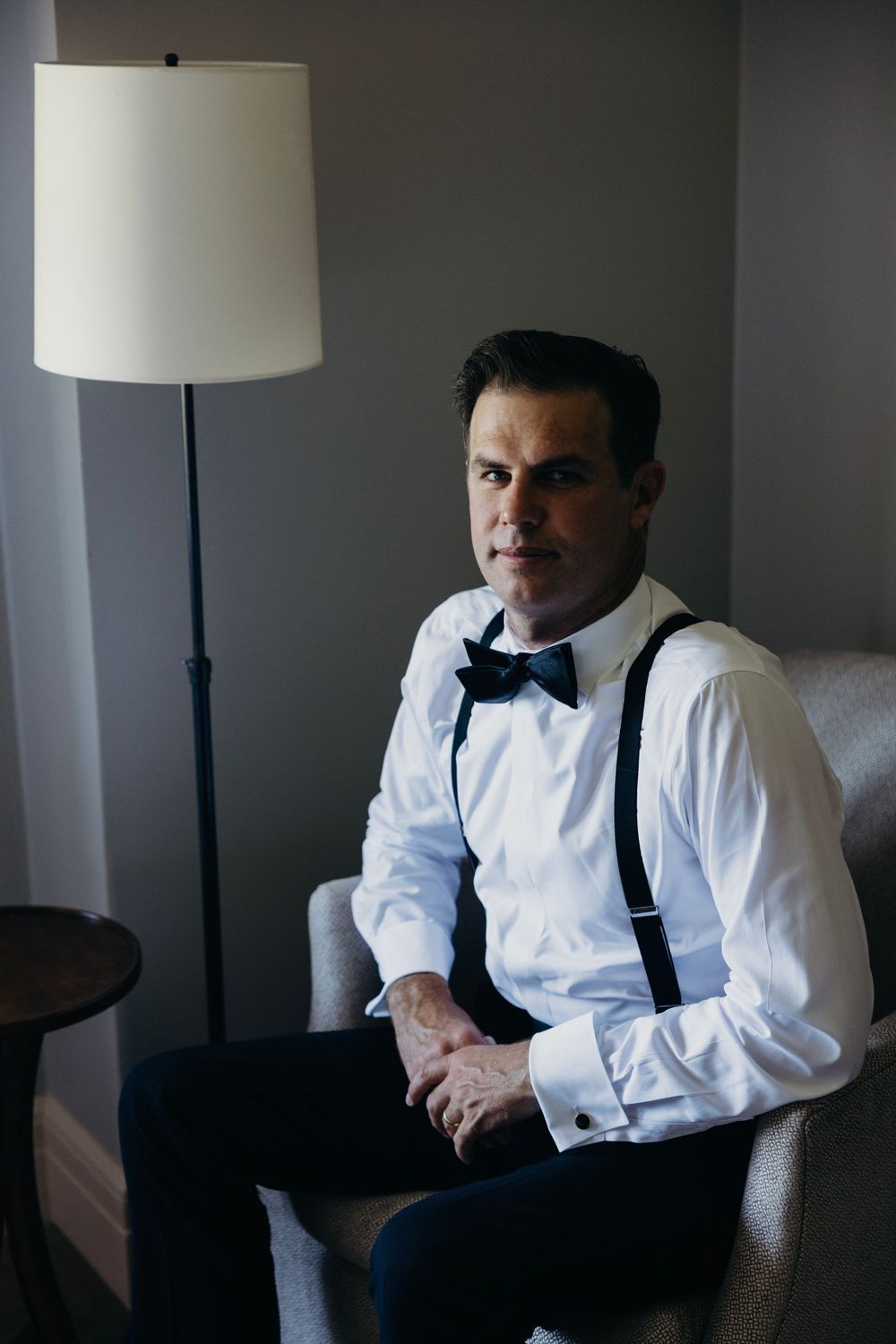 Person in formal attire seated in moody lighting with lamp.