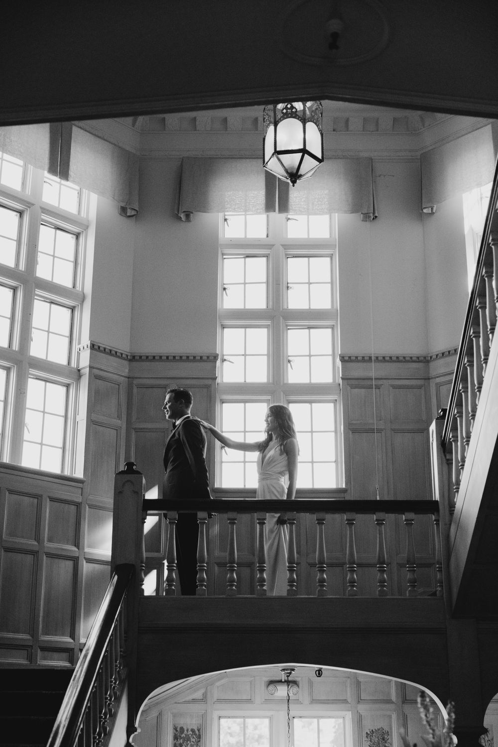 Black and white photo of a couple meeting on a grand staircase landing with dramatic window lighting.