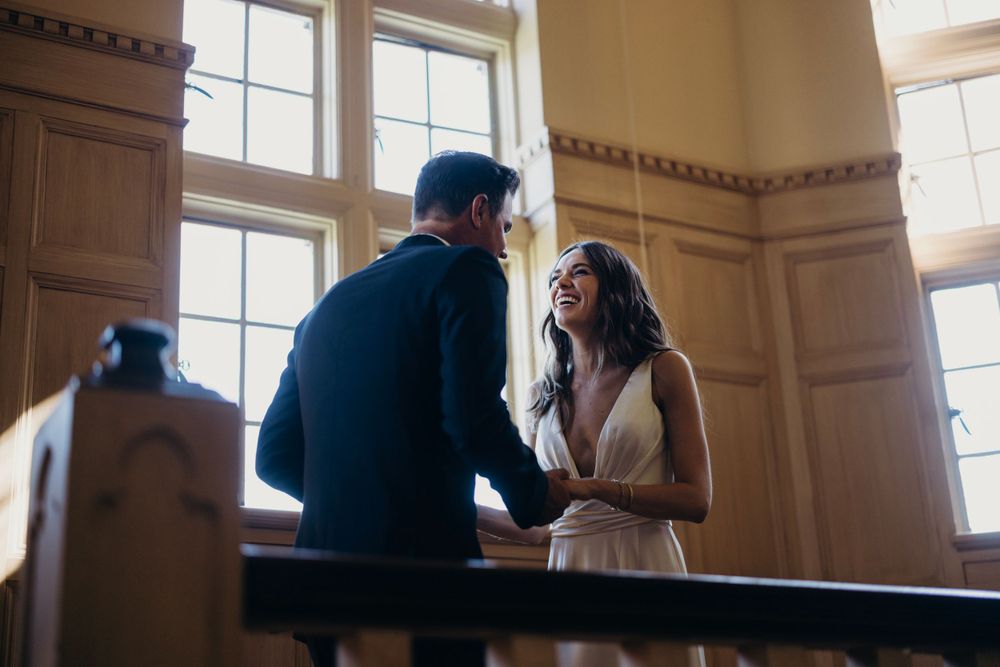Intimate moment between newlyweds in front of tall windows with natural light streaming in.