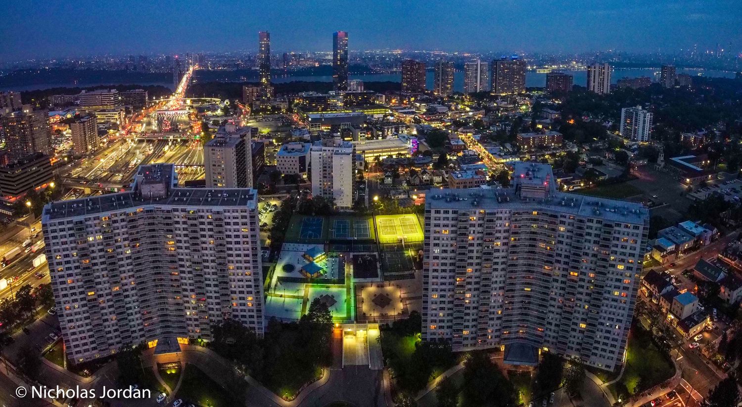 Night cityscape with illuminated high-rise buildings and tennis courts glowing in green.