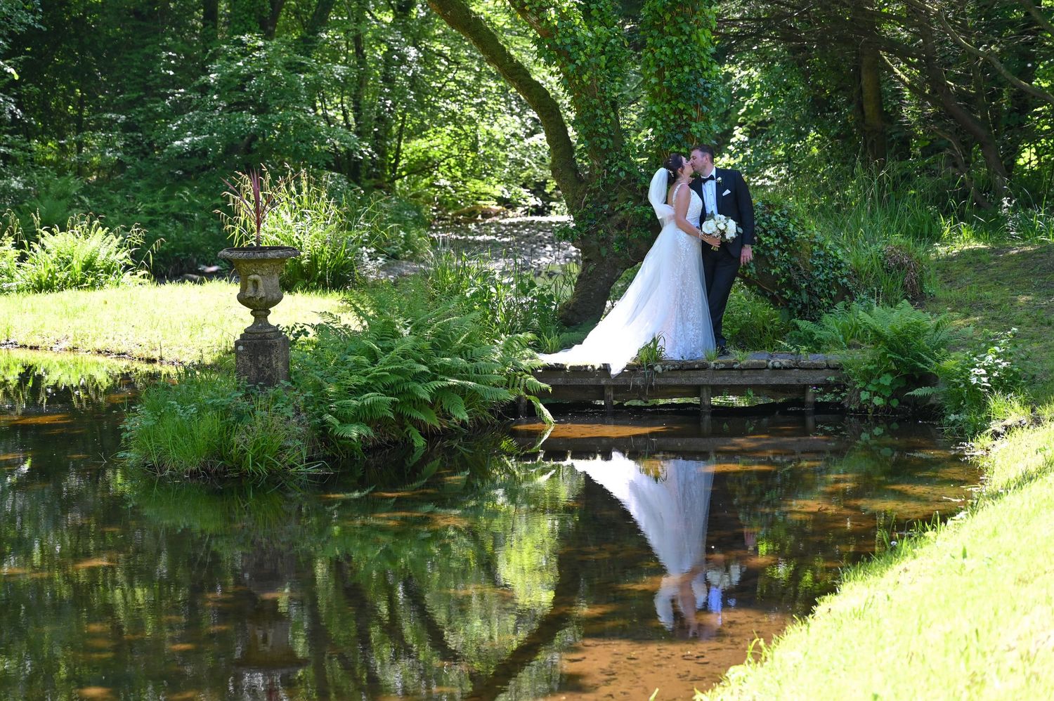 A couple in wedding attire poses on a wooden bridge over a reflective pond in a lush garden setting.