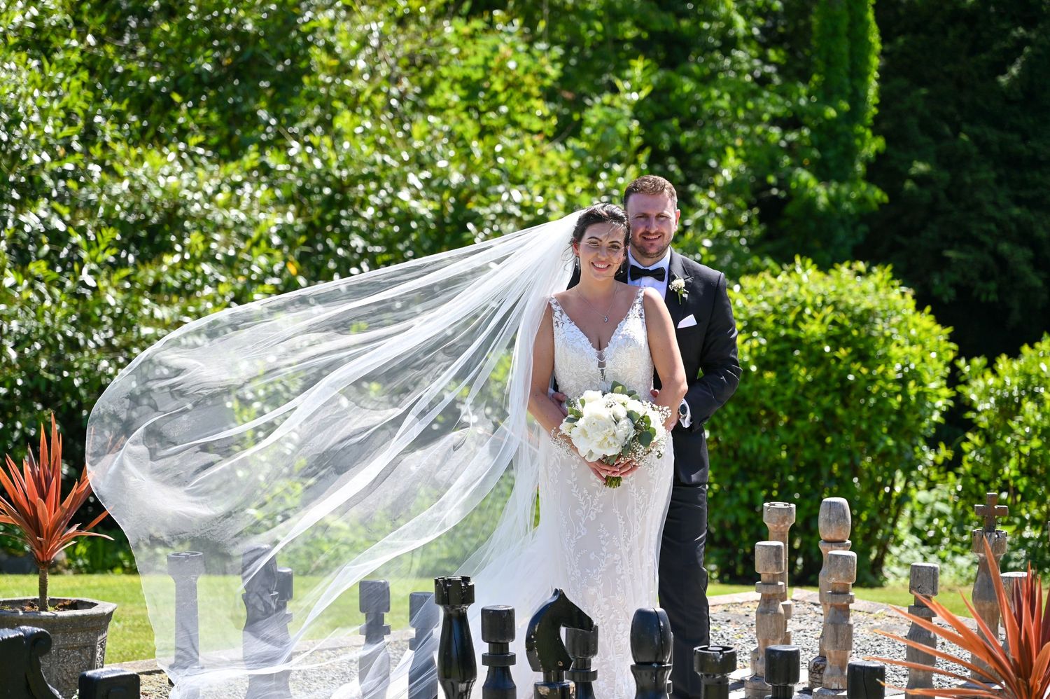 Bride's veil flows dramatically in the wind as couple poses in garden with decorative pillars.