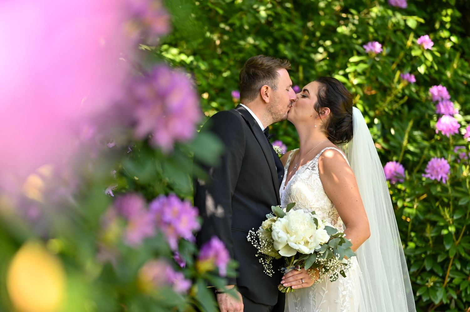 Wedding couple shares a kiss among pink flowers with white bouquet in hand.