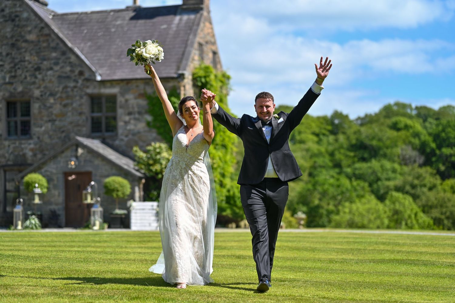 Joyful newlyweds celebrate with raised arms on mansion grounds under blue sky.