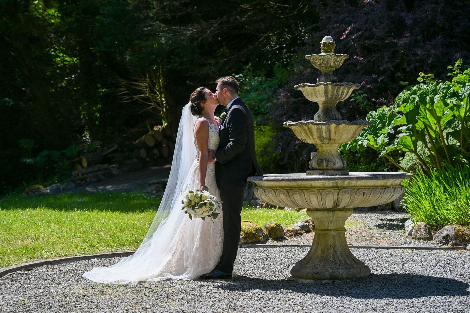 Newlyweds share an intimate moment beside a decorative stone fountain in a garden setting.