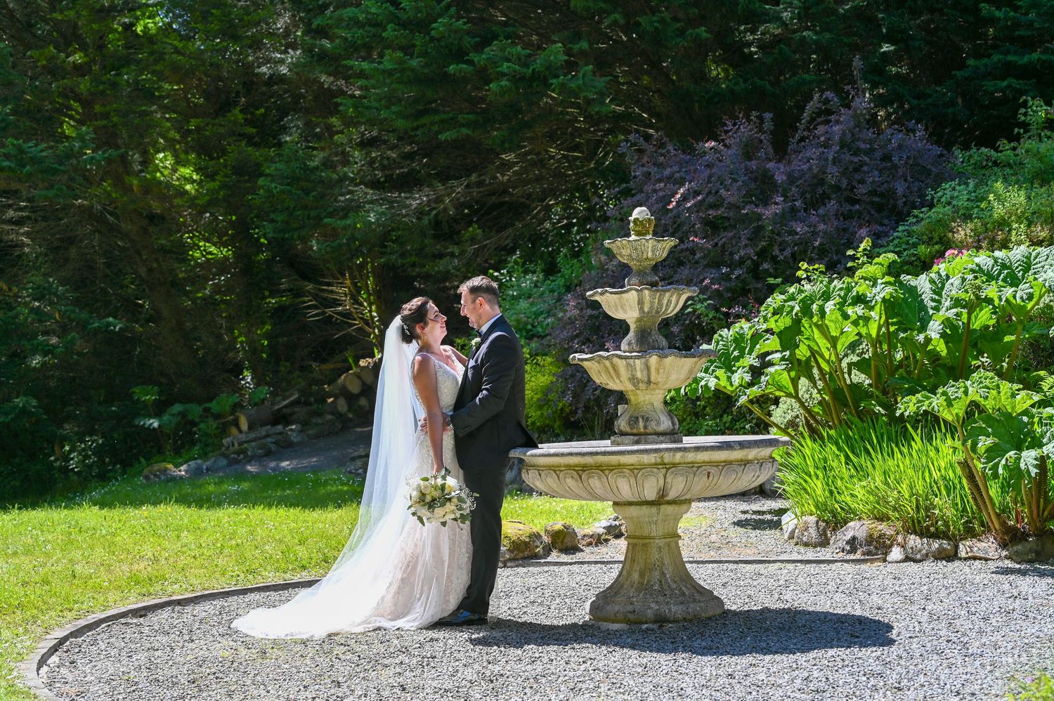 Wedding couple stands beside an ornate tiered fountain in a garden with gravel path.