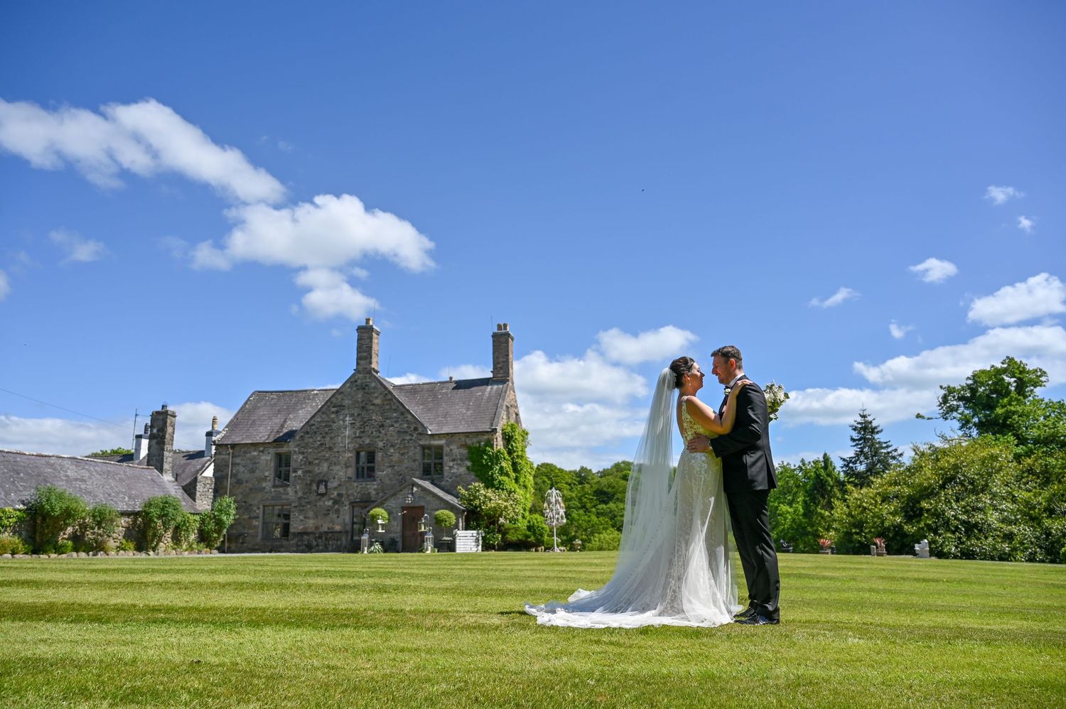 Newlyweds stand on green lawn with historic stone building in background under blue sky.