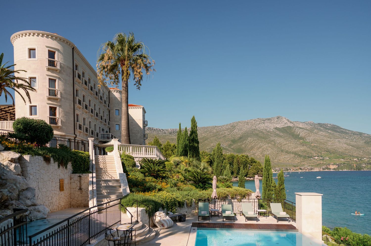 Luxury Mediterranean hotel with swimming pool overlooking coastal mountains and sea.