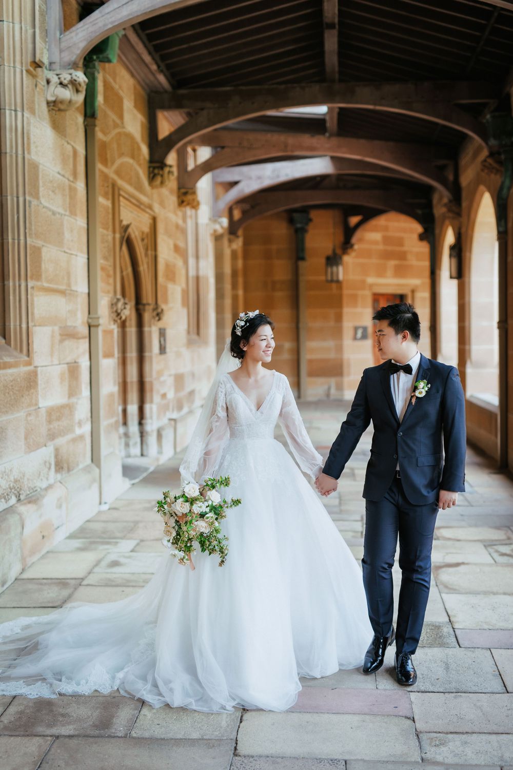 Bride and groom walking hand in hand through arched sandstone corridor with floral bouquet.