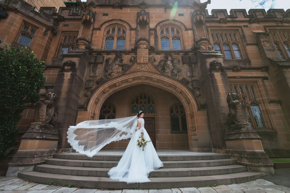 Bride in white gown with flowing veil standing on steps of ornate historic stone building.
