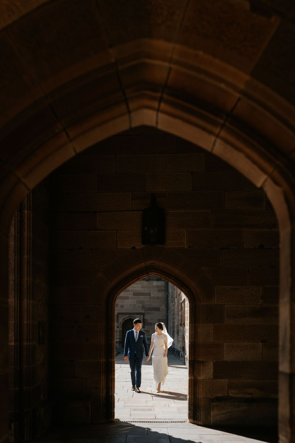 Wedding couple walking hand in hand through gothic stone archway in warm sunlight.