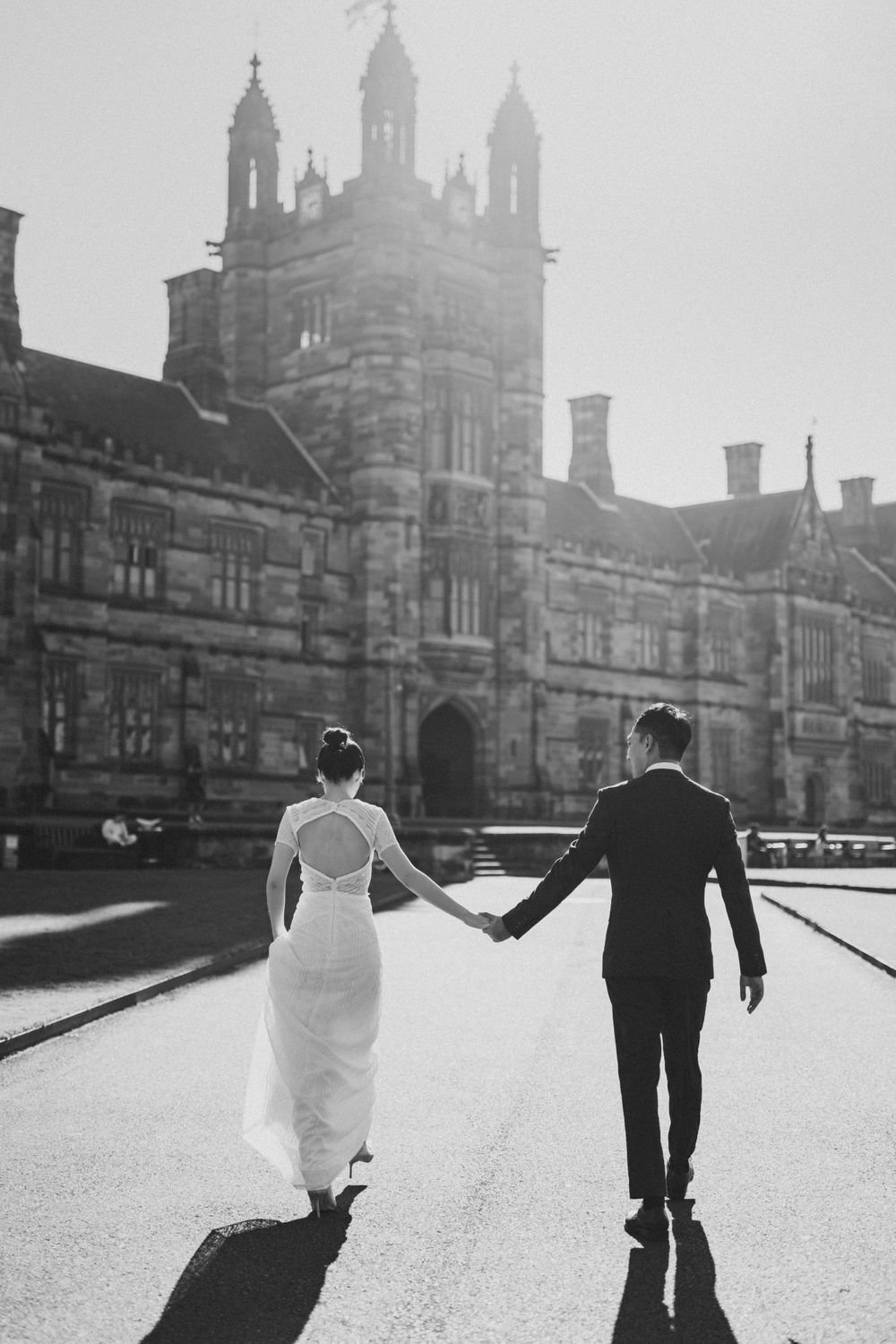 Black and white photo of bride and groom holding hands walking toward historic stone building.