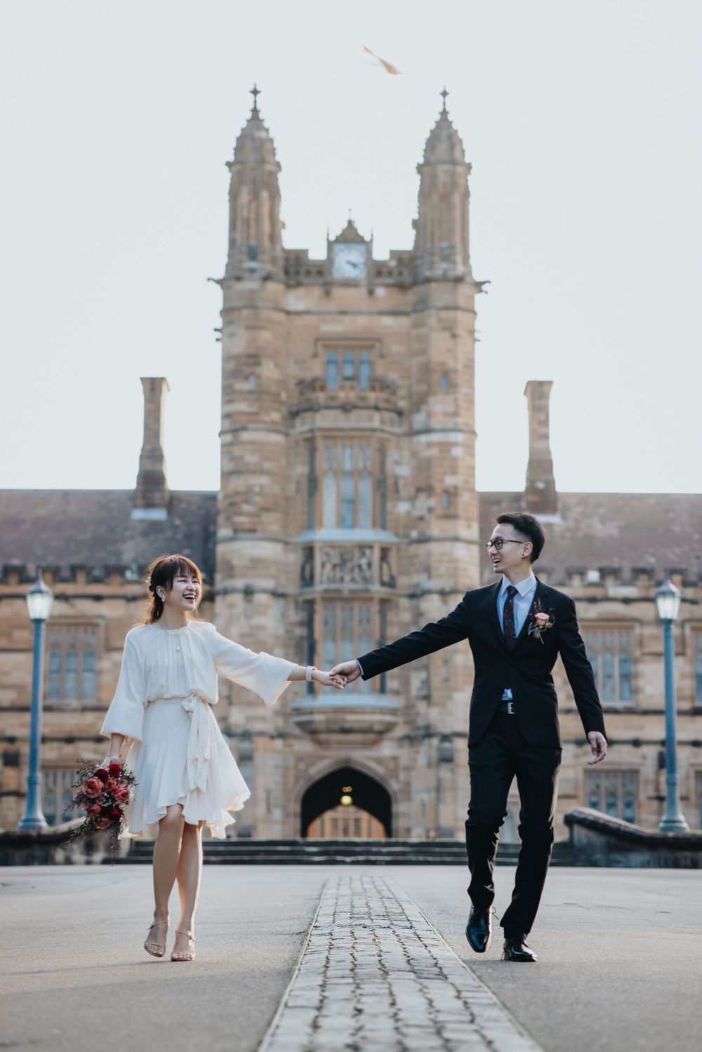 Smiling bride in white dress holding bouquet with groom before historic sandstone university building.