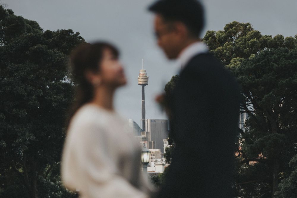 Blurred couple in wedding attire with Sydney Tower visible among trees in background.