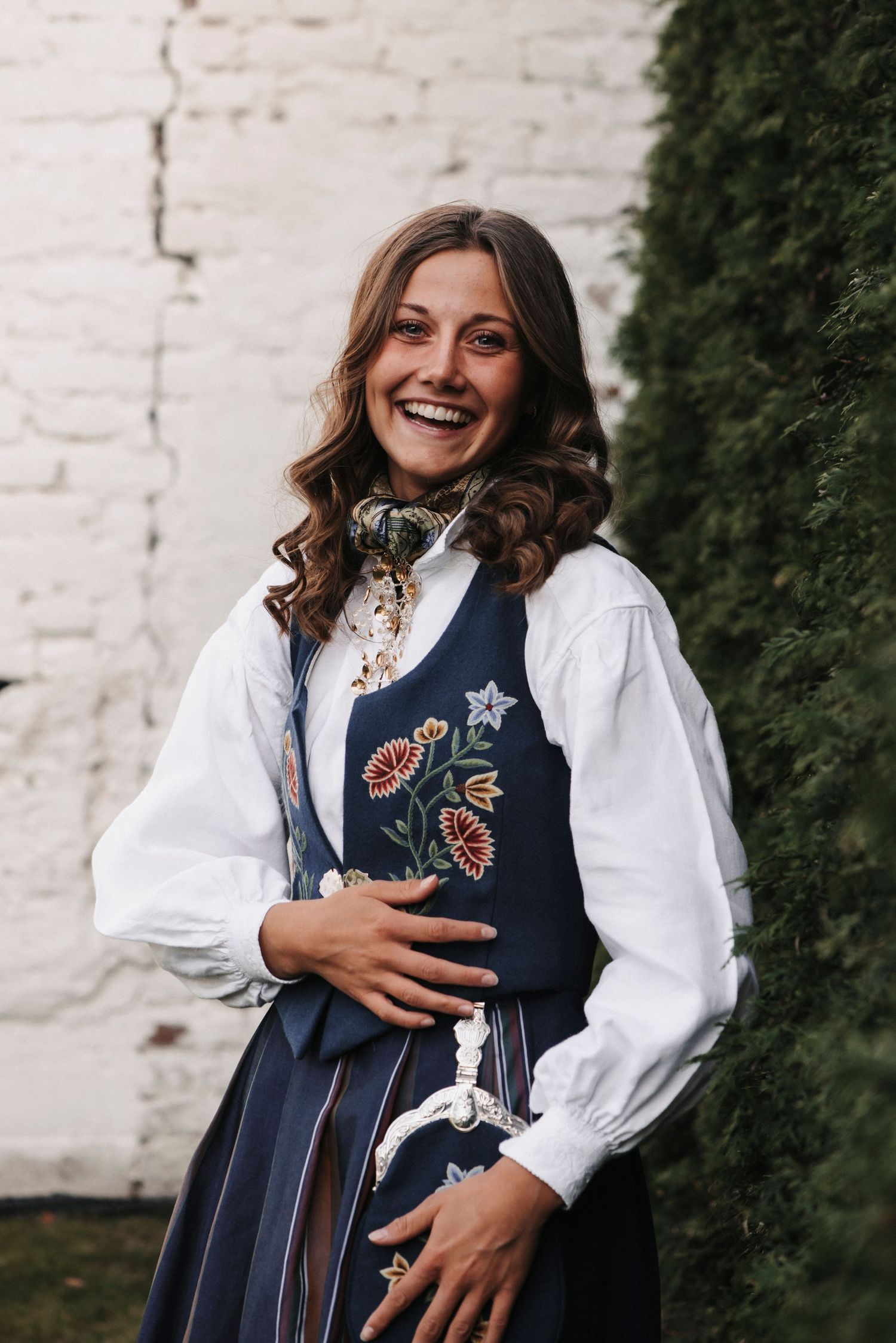 A woman wearing a Norwegian traditional dress with embroidered floral details smiles against a white wall.