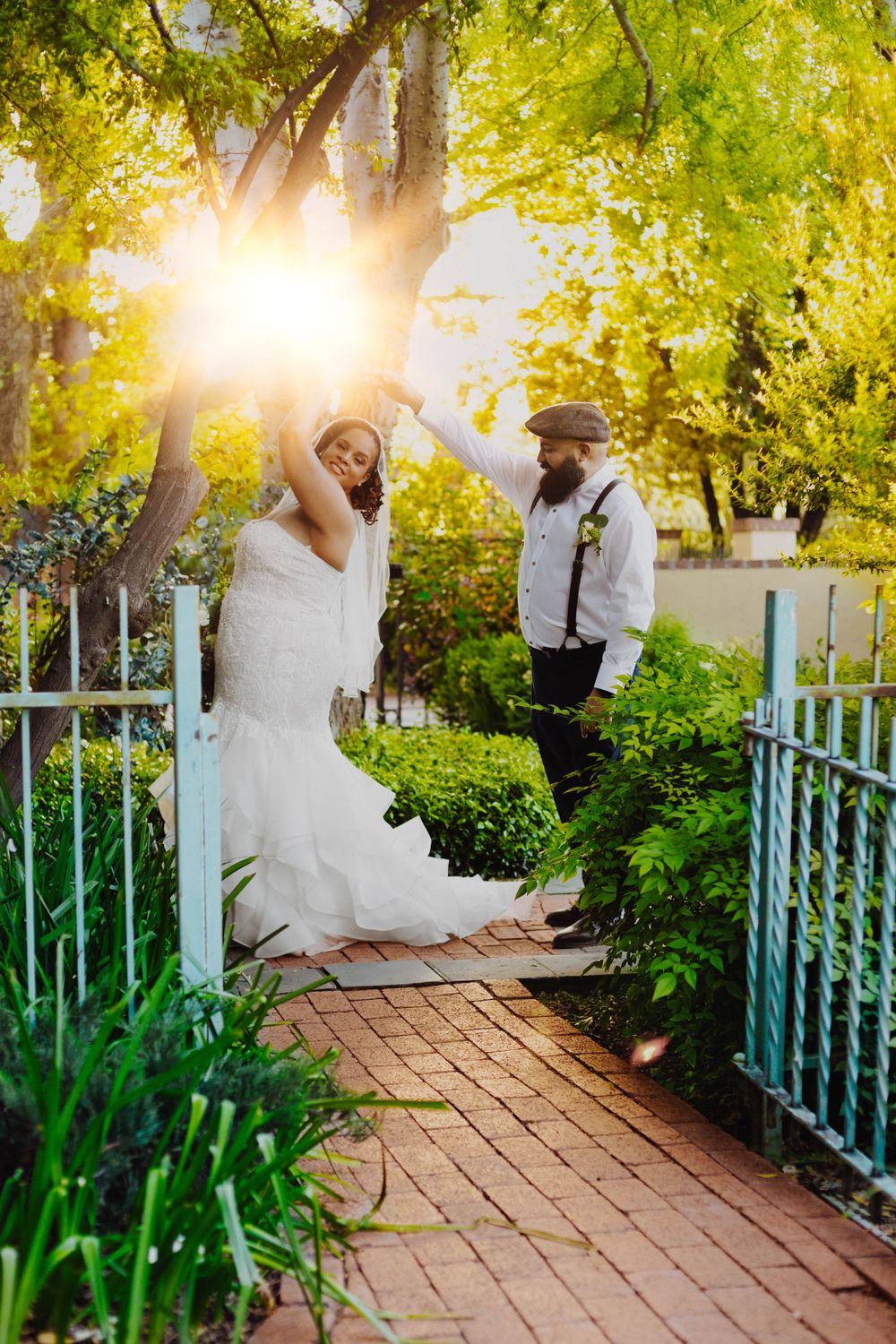 Wedding couple poses on brick path with sunburst through green garden trees.