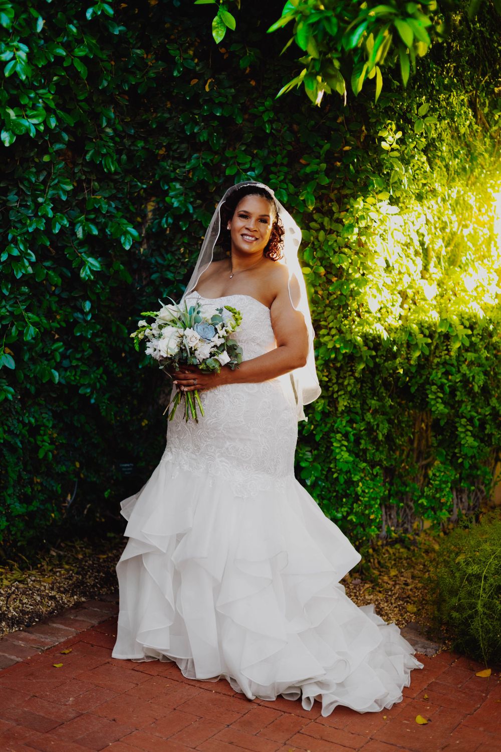 Bride in strapless mermaid gown holds bouquet while posing against green foliage.
