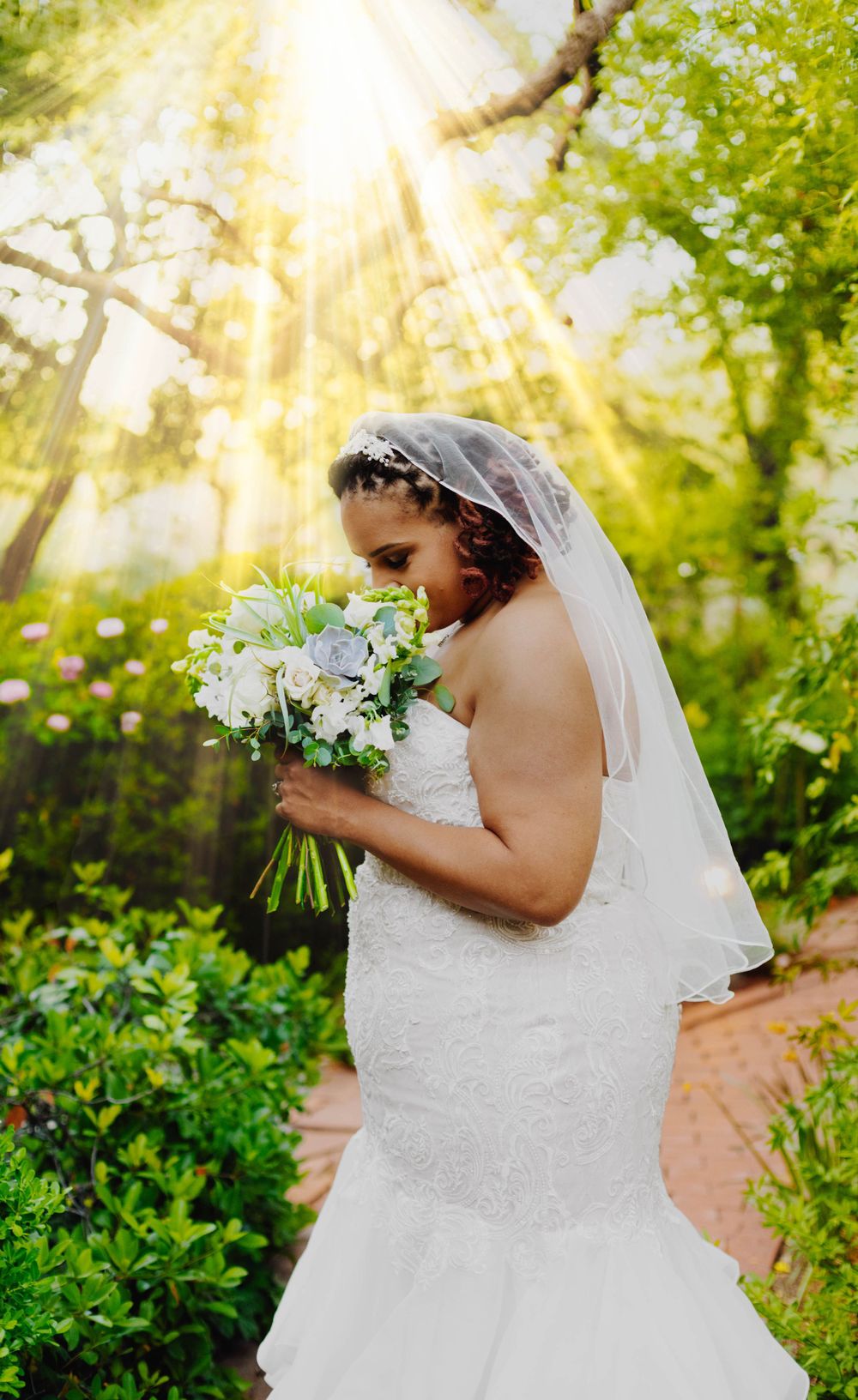 Bride with bouquet of white flowers poses in garden with dramatic sunlight streaming through trees.