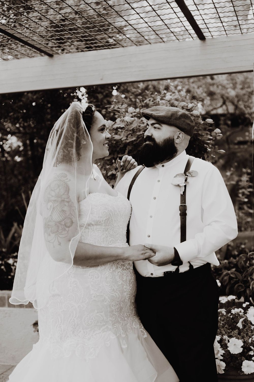 A black and white portrait of a wedding couple posing under a latticed garden structure.