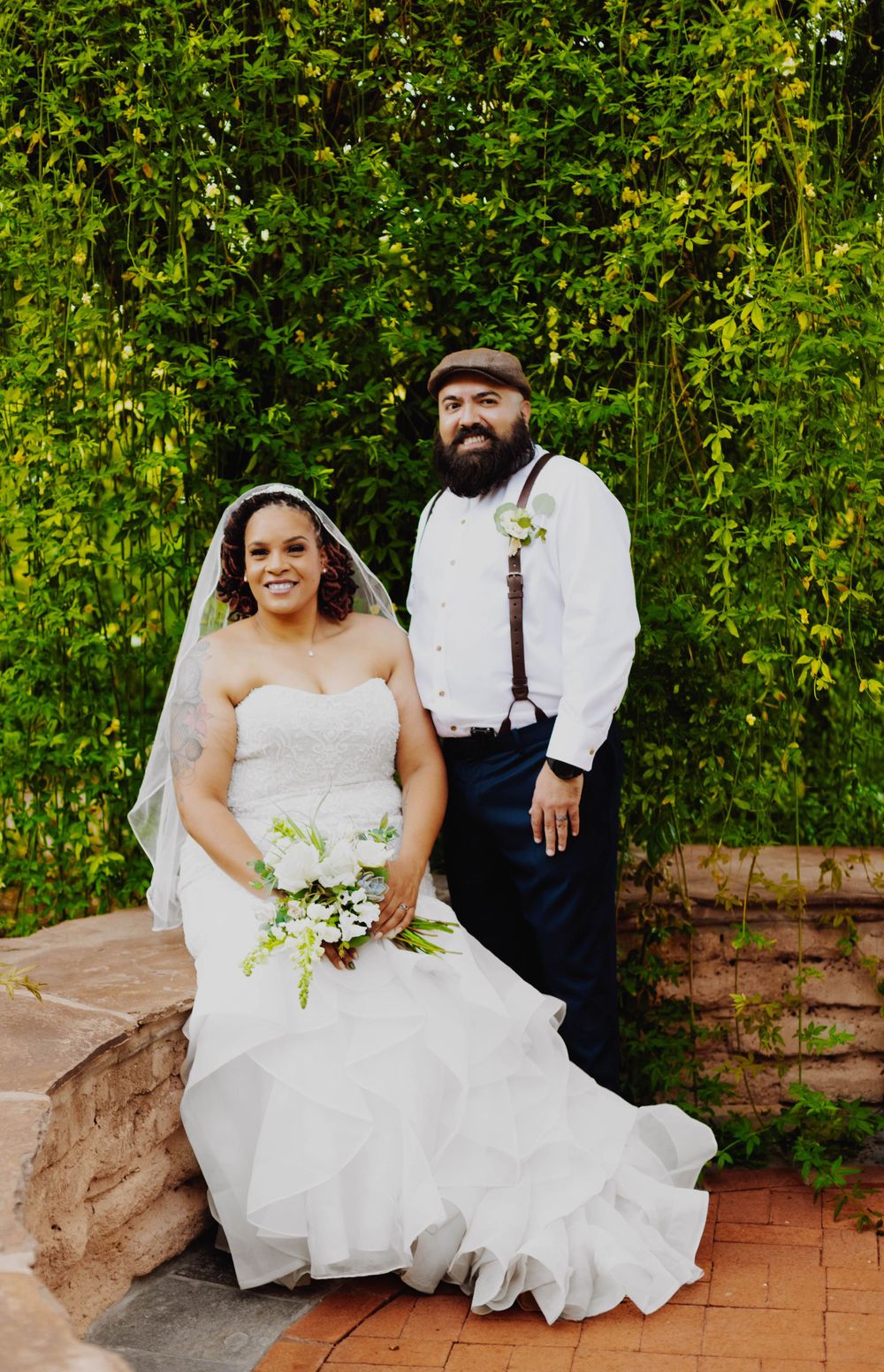 Newlyweds pose together against a wall of green foliage on brick steps.
