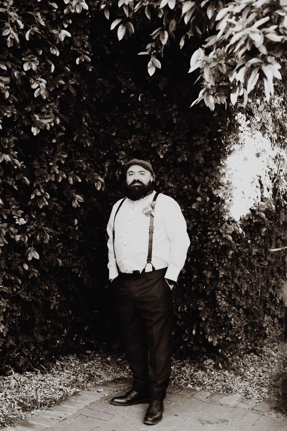 Black and white portrait of groom in suspenders standing against leafy backdrop.
