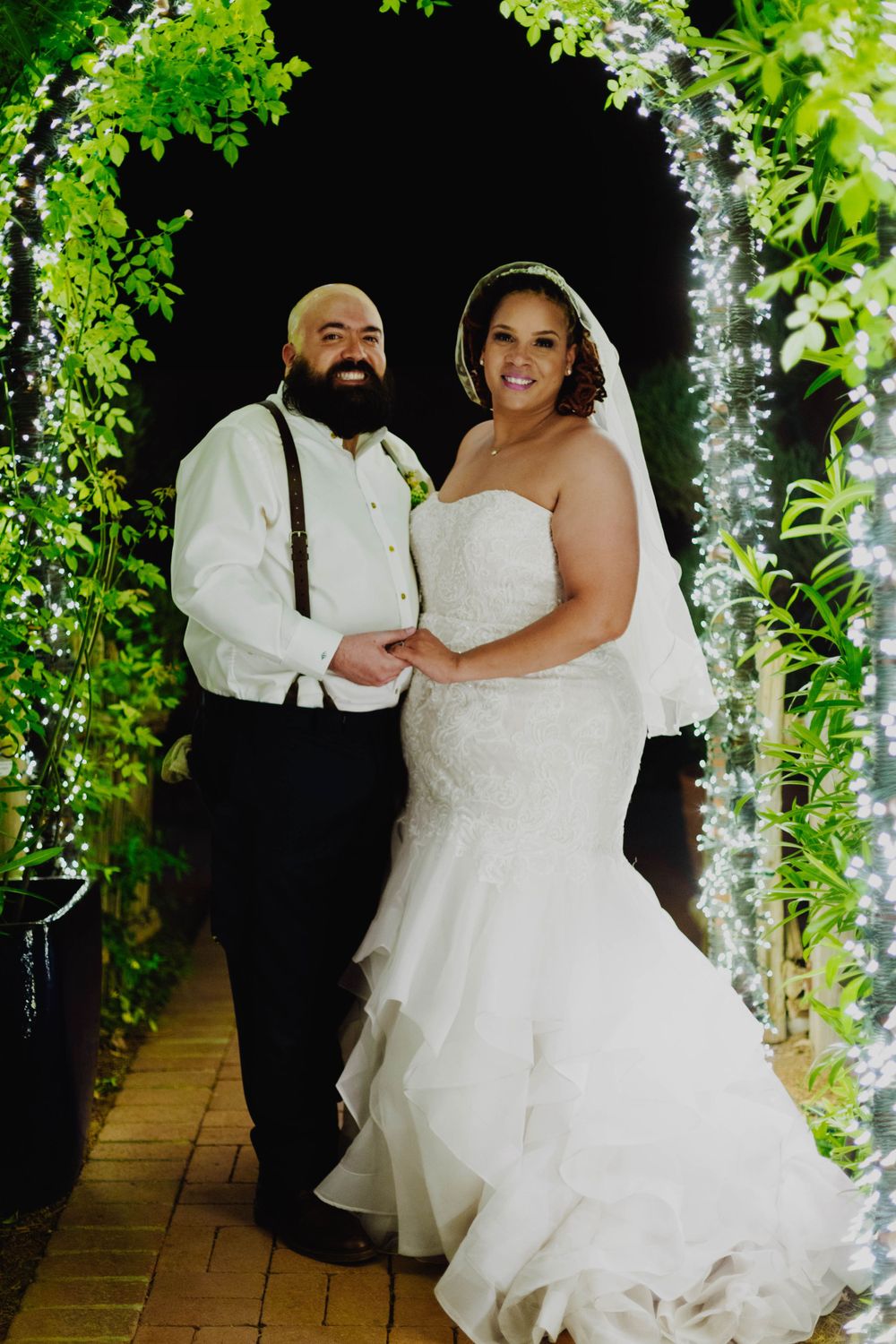 Wedding portrait under illuminated garden archway with greenery and twinkling lights.