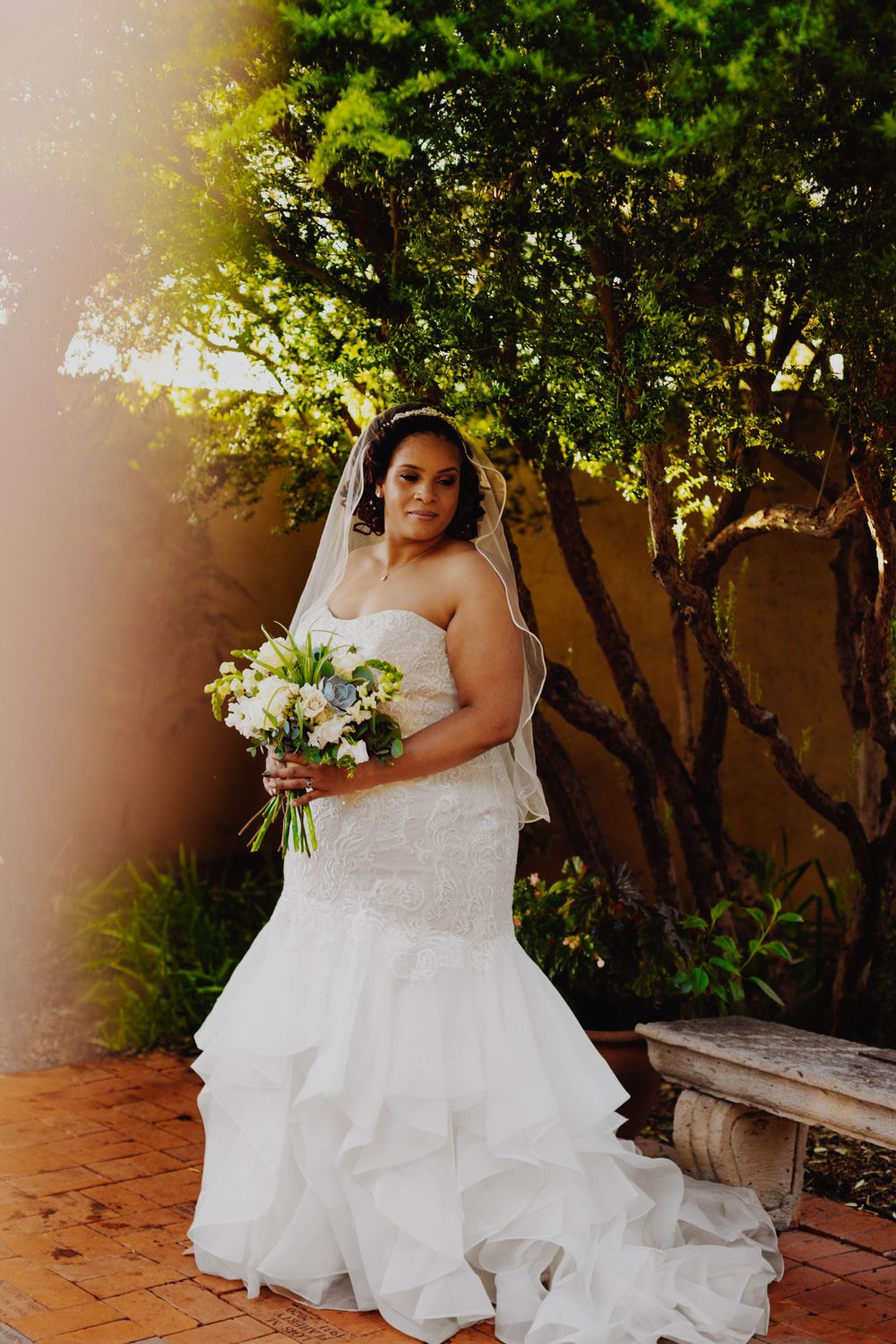A bride in a flowing white dress holds a bouquet against a lush garden backdrop with sunlit trees.