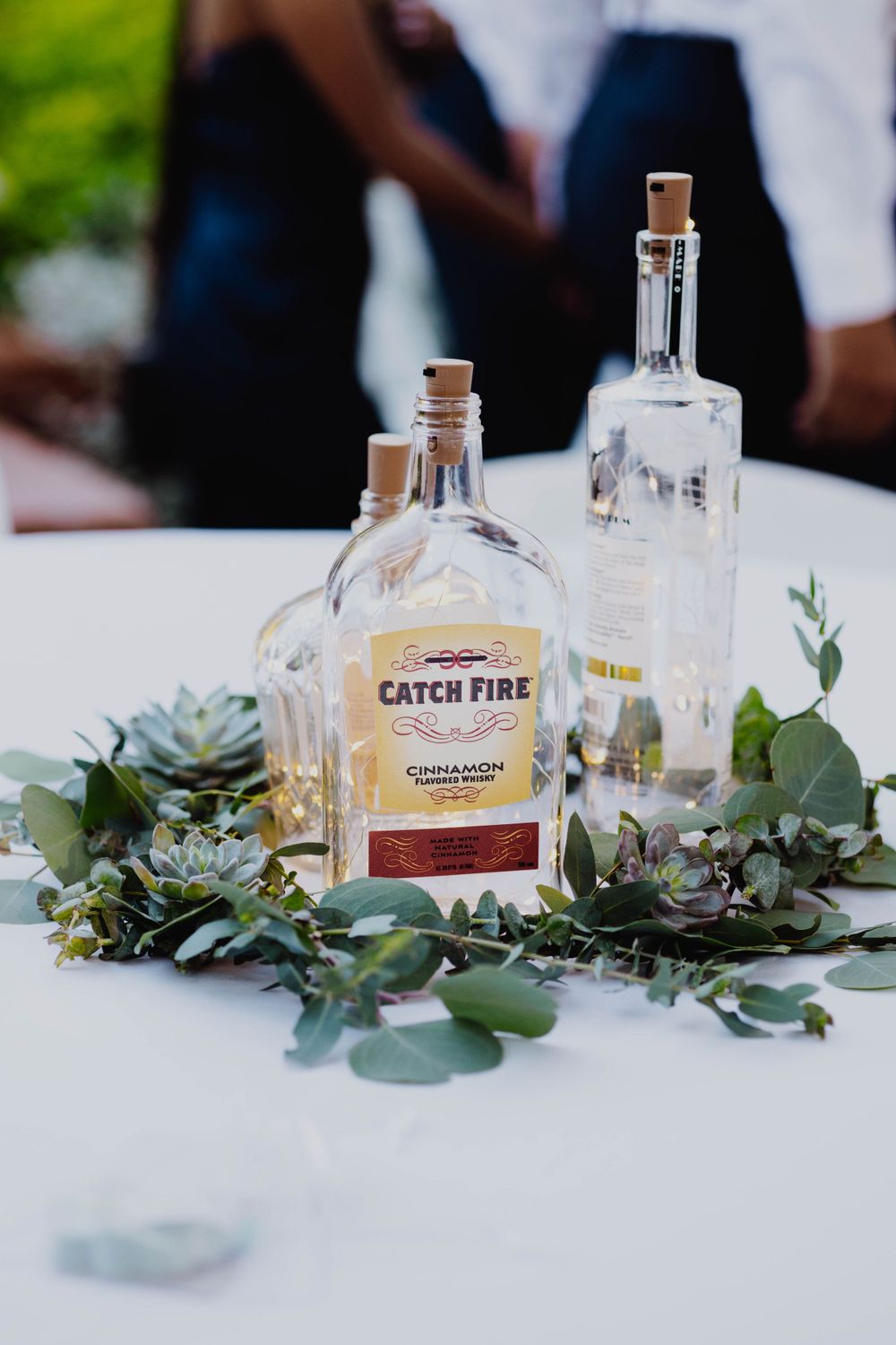Glass bottles with eucalyptus leaves arranged as wedding table decor on a white tablecloth.