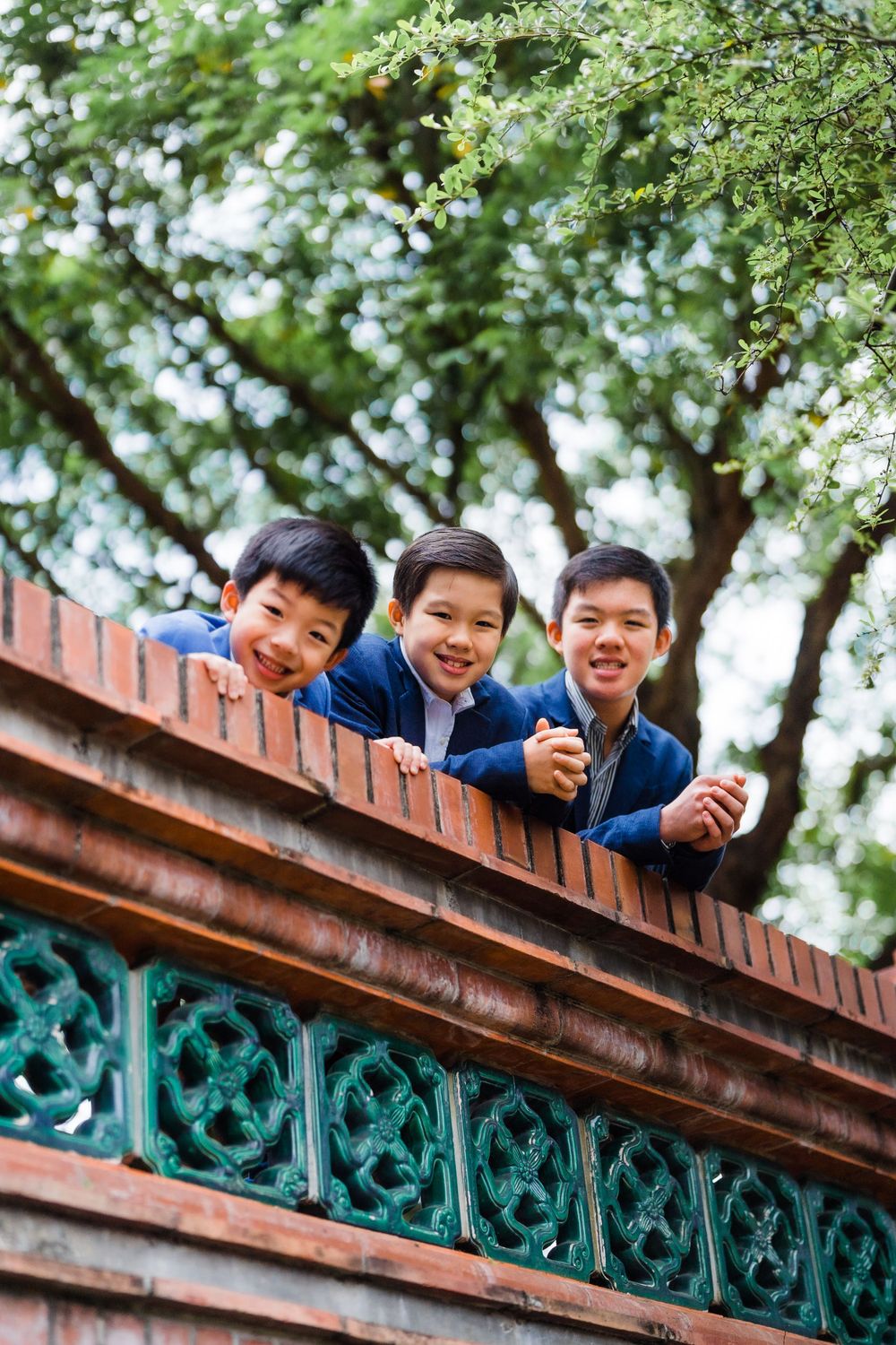 Three boys in blue jackets peek over a decorative brick wall with ornate green details.